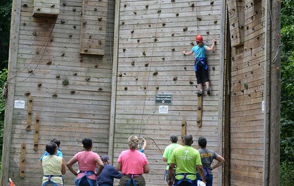 a group of people look on as a participant climbs a wall at the obstacle course